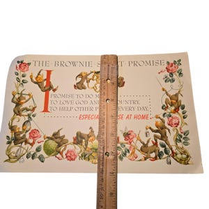 May include: Vintage print featuring whimsical illustrations of brown gnomes and pink roses. The print includes text that reads "THE BROWNIE ST-PROMISE" and other text. A wooden ruler is placed vertically on the print, measuring approximately 8 inches.