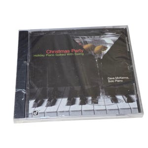 Puede incluir: Una caja de CD negra con la inscripción "Christmas Party" y "Holiday Piano Spiked With Swing". La portada incluye una copa de Martini con aceitunas y un teclado de piano. El artista es Dave McKenna.