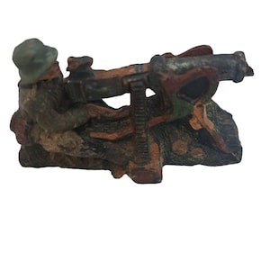 May include: A small, hand-painted, toy soldier figurine holding a machine gun. The soldier is wearing a green hat and brown uniform. The machine gun is painted in shades of brown, green, and red.