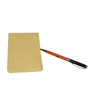 May include: A light yellow notepad with a pen. The pen has an orange barrel with black accents and a silver clip. The notepad has a rounded rectangle shape and a small embossed design in the lower left corner.