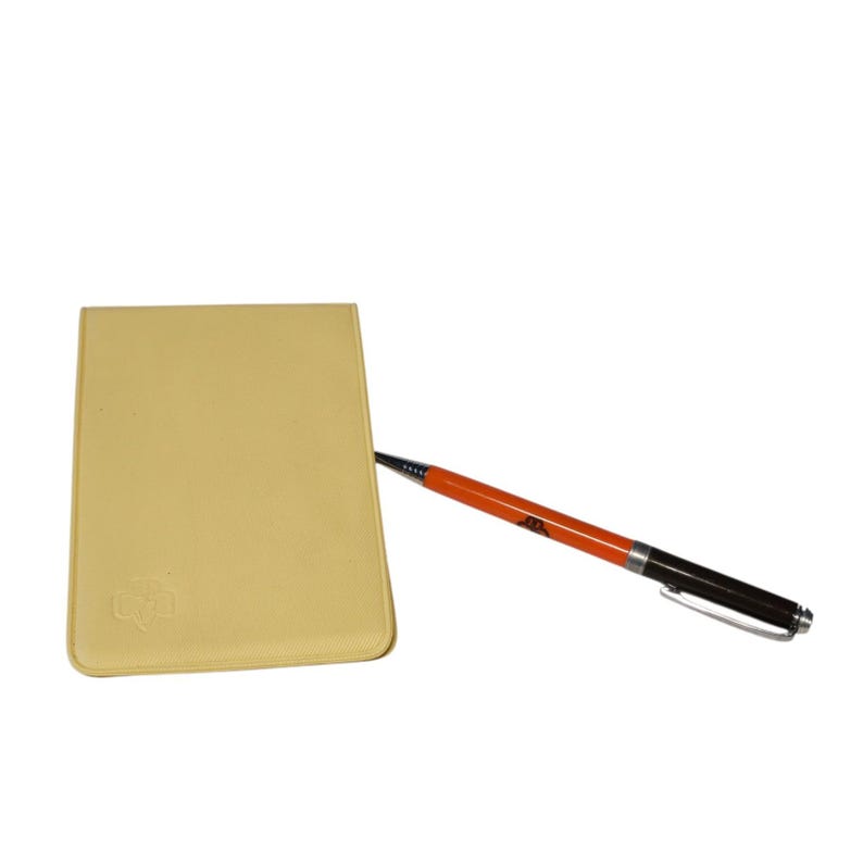 May include: A light yellow notepad with a pen. The pen has an orange barrel with black accents and a silver clip. The notepad has a rounded rectangle shape and a small embossed design in the lower left corner.