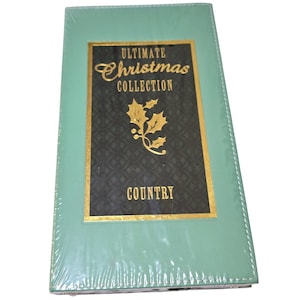 Puede incluir: Un artículo rectangular verde menta envuelto en plástico transparente. La parte delantera presenta una etiqueta negra y dorada con las palabras "ULTIMATE Christmas COLLECTION COUNTRY" y un diseño de rama de acebo dorada.