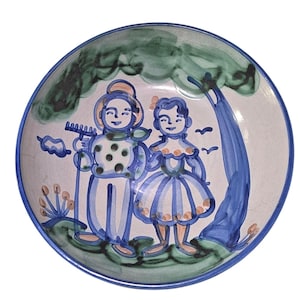 Puede incluir: Un cuenco de cerámica pintado a mano con un diseño caprichoso. El cuenco presenta dos figuras bajo un árbol, pintadas en azul, verde y naranja. El borde es azul y el fondo es blanco. El cuenco es una pieza decorativa.