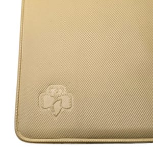 May include: A beige, rectangular item with a textured surface and rounded corners. The item features a debossed emblem in the lower left corner. The emblem is a stylized floral design. The item is a light tan color.