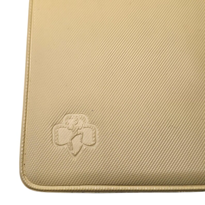 May include: A beige, rectangular item with a textured surface and rounded corners. The item features a debossed emblem in the lower left corner. The emblem is a stylized floral design. The item is a light tan color.
