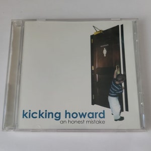 Puede incluir: Una caja de CD con fondo blanco y una foto de un joven empujando una puerta con la etiqueta "women". El texto "kicking howard an honest mistake" está impreso en azul en la parte delantera de la caja.