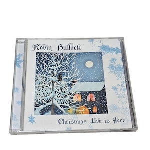 May include: A CD case with the album "Christmas Eve is Here" by Robin Bullock. The cover art shows a snowy winter scene with a house, a full moon, and a tree. The case is clear with blue snowflake accents.
