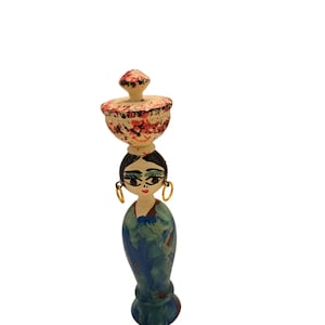 May include: A hand-painted wooden figurine of a woman carrying a bowl on her head. The figurine has a blue and green dress, black hair, gold hoop earrings, and a painted face. The bowl is beige with red floral accents.
