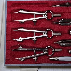 May include: A set of silver drafting compasses and tools arranged in a red velvet-lined case. The tools include various compasses, dividers, and a pencil holder, all with a metallic finish. The case has a gray border.