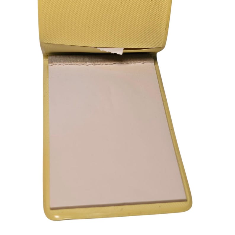 May include: A small, open, yellow notepad with a textured cover and a stack of blank, light pink pages. The pages are held together at the top with a rough edge. The notepad is open, revealing the pages.