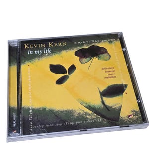 May include: A CD case featuring the album "in my life" by Kevin Kern. The cover art has a yellow and green background with a floral design. Text includes "delicately layered piano melodies" and "REALMUSIC".