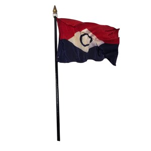 Pode incluir: Uma bandeira retangular presa a um mastro preto com um remate de cor dourada. A bandeira apresenta uma faixa horizontal vermelha no topo e uma azul-escura na parte inferior, com um triângulo branco contendo um círculo azul-escuro.