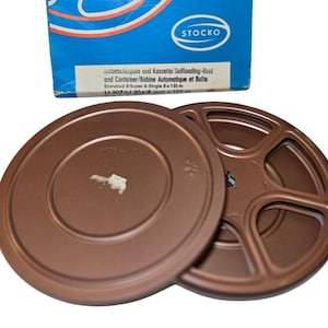 Könnte beinhalten: Ein Vintage-Filmrollen- und Behälter-Set. Die Rollen sind warmbraun, mit einem passenden Behälterdeckel. Die blaue Box im Hintergrund trägt den Markennamen "STOCKO" und Text auf Deutsch und Französisch.