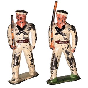 May include: Two vintage toy soldier figurines. Each figure is painted in off-white with black and brown accents, wearing a sailor hat and holding a staff. The figures have a distressed, aged appearance.