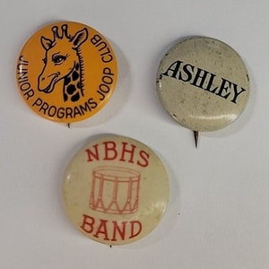 Könnte beinhalten: Drei Vintage-Ansteckbuttons. Einer ist gelb mit einer Giraffe und dem Text "Junior Programs Joop Club". Ein weiterer ist cremefarben mit dem Namen "Ashley". Der dritte ist cremefarben mit einer Trommelgrafik und dem Text "NBHS Band".
