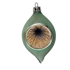 May include: A vintage Christmas ornament in a teardrop shape. The ornament has a pale green exterior with a silver-colored top and a circular, textured design in the center. The ornament is suspended by a small metal loop.