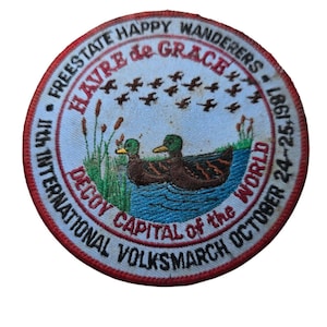 May include: A white circular patch with red trim, featuring a scene of two ducks swimming in a blue pond. The text reads "Freestate Happy Wanderers - 18th International Decoy Capital of the World - Volksmarch October 24-25, 1981" and "Havre de Grace".