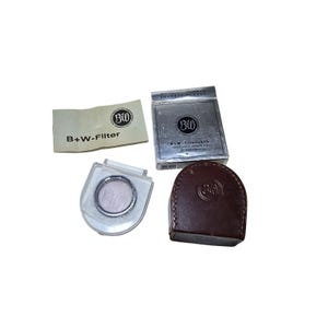 Puede incluir: Un conjunto de accesorios de filtro B+W, que incluye un filtro, una funda, una caja y un papel. El filtro está en una funda de plástico transparente. La caja y la funda son plateadas y marrones. El papel tiene el texto "B+W-Filter".