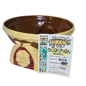 May include: A ceramic Scorpion Bowl with a brown interior and a cream-colored exterior with brown and maroon accents. The bowl has a unique design with a carved face and the text "NAME YOUR POISON". A tag reads "OFFICIAL HUKILAU 2005 SCORPION BOWL".