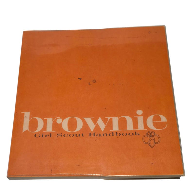 May include: An orange Brownie Girl Scout Handbook. The cover features the word "brownie" in a large, off-white font, with "Girl Scout Handbook" below. The Girl Scout trefoil logo is in the lower right corner.
