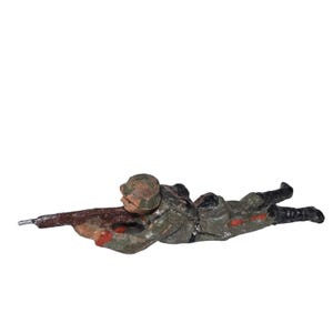 May include: A vintage toy soldier in a prone position, holding a rifle. The figure is painted in shades of green, brown, and black, with red accents. The toy soldier is lying down, ready for action.