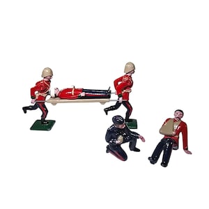 Puede incluir: Un conjunto de soldaditos de juguete que representan una escena médica. Tres soldados con uniformes rojos llevan una camilla con un soldado herido. Otros dos soldados atienden las heridas. Las figuras están sobre bases verdes.