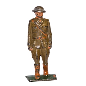 May include: A vintage cast iron toy soldier figurine. The figure is painted in olive green with orange accents on the boots and strap. The soldier wears a helmet and stands on a green rectangular base. The figurine is a collectible item.