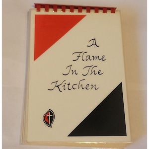 May include: A white cookbook with a red and black diagonal design. The title is "A Flame In The Kitchen". A small logo with a red flame and a cross is in the bottom left corner.