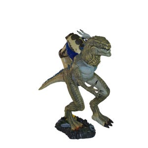 Könnte beinhalten: Detaillierte Actionfigur einer reptilienartigen Kreatur mit humanoider Form. Die Figur ist hauptsächlich grün und beige, mit blauen Akzenten. Sie steht auf einem felsigen Sockel und hat scharfe Krallen und Zähne. Die Figur ist etwa 25 cm groß.