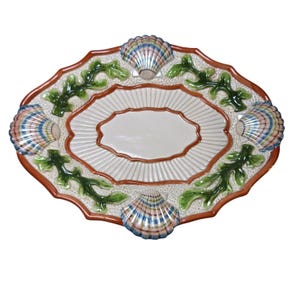 May include: An oval ceramic serving platter featuring a central cream-colored basin with radiating fluted lines. The rim is decorated with four raised, multi-colored scallop shells and green seaweed motifs against a textured off-white background.