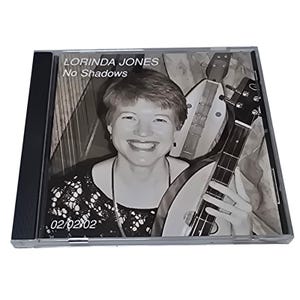 Puede incluir: Una caja de CD con el álbum "No Shadows" de Lorinda Jones. La portada es una foto en blanco y negro de una mujer sonriente sosteniendo un instrumento de cuerda. El texto "LORINDA JONES" y "No Shadows" son visibles.