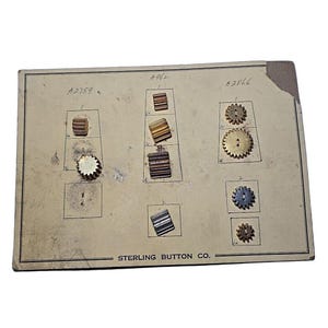 May include: Aged cardboard display featuring various small metal gears and components. The display is labelled with numbers and the text "STERLING BUTTON CO." The gears are in shades of gold, silver, and bronze, showcasing intricate designs.