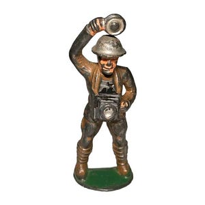 May include: A vintage toy soldier figurine made of metal, painted in shades of gray and brown. The soldier is wearing a helmet and holding a camera with a flash. The figurine is standing on a green base.