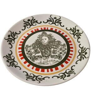 May include: A white ceramic plate with a central portrait of a historical figure, surrounded by red, black, and yellow concentric circles. The plate features decorative black floral patterns around the rim. The portrait has text below it.