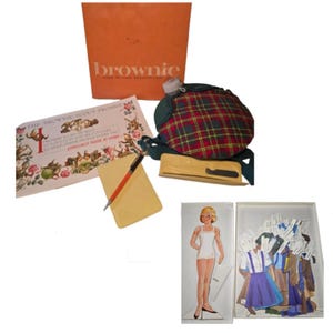 May include: Vintage Brownie Girl Scout Handbook and accessories. Includes an orange box with the word "brownie", a plaid canteen, paper doll with outfits, a notebook, pen, and a booklet with text.