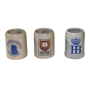 May include: Three small, cylindrical ceramic beer steins. The steins are off-white with various logos and text in blue and orange. One stein has a blue silhouette and the text "THOMASBRAU MUNCHEN". Another has a logo and the text "SPATENBRAU MUNCHEN". The third has a crown and the letters "HB".