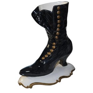 May include: A glossy black ceramic boot-shaped vase featuring a row of raised gold-toned buttons along the side. The boot rests on a white, scalloped base with a gold-painted edge, and the interior of the boot is finished in a smooth white glaze.