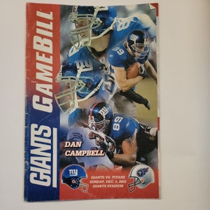 May include: A blue and red game day program for the New York Giants football team. The program features a photo of Dan Campbell, a player for the Giants, and the text "GIANTS GAMEBILL" and "GIANTS VS. TITANS SUNDAY, DEC. 1, 2002 GIANTS STADIUM".