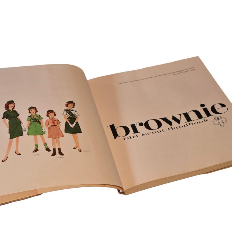 May include: Open book with the title "Brownie Girl Scout Handbook" in black text. The left page features illustrations of four girls in Brownie uniforms. The book has a light tan cover and pages.
