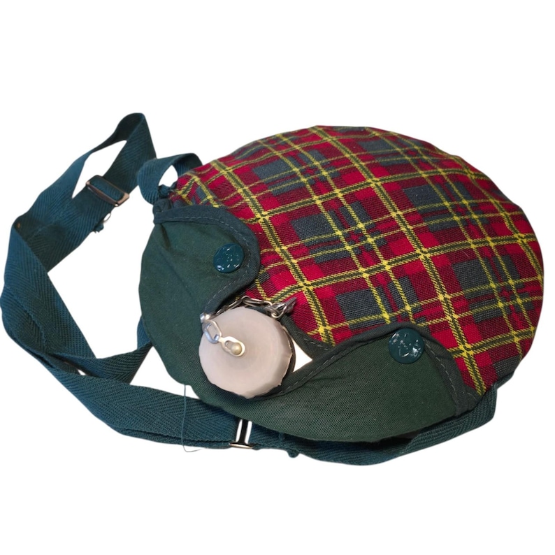 May include: A vintage canteen with a plaid pattern in red, yellow, and black. The canteen has a green fabric base and strap, with a metal clasp and a small, round, light-colored stopper. The canteen is designed to be worn over the shoulder.