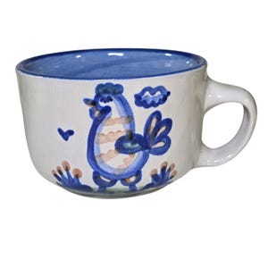 Puede incluir: Taza de cerámica blanca con interior y asa azules. La taza presenta un diseño pintado a mano de un gallo azul con detalles en melocotón y azul. Un pequeño pájaro y una nube azules también están pintados en la taza.