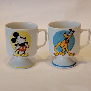 Peut inclure: Deux tasses en céramique blanche avec un piédestal. Une tasse présente une image de dessin animé de Mickey Mouse en short rouge et chaussures jaunes. L'autre tasse présente une image de dessin animé de Pluto le chien en jaune et marron. Les deux tasses ont une bordure bleue et jaune autour de la base.