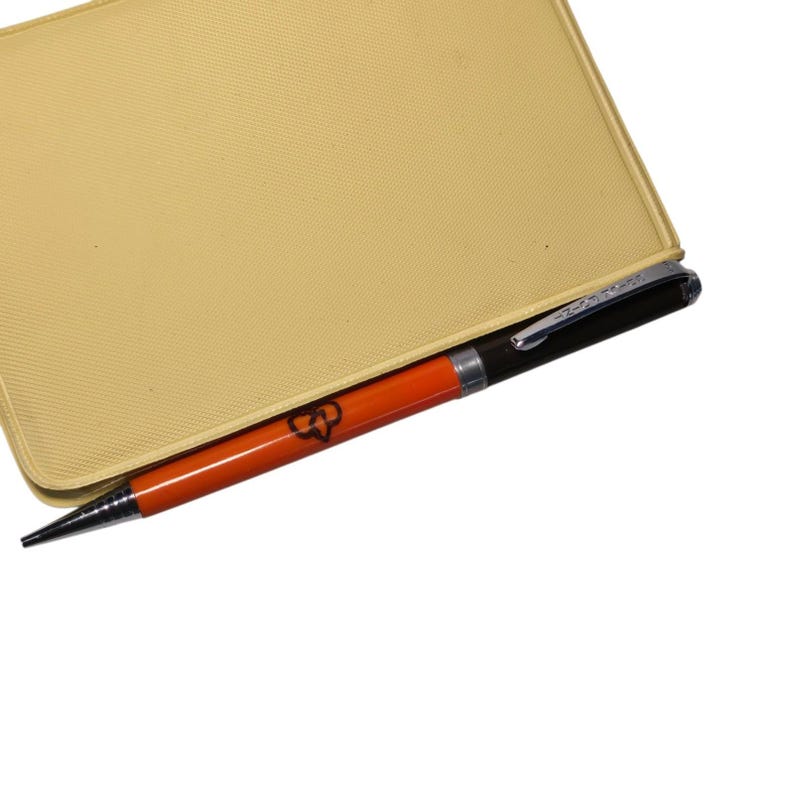 May include: A gold-colored notebook with a textured surface. An orange and black pen with a silver tip is inserted into the notebook. The pen has a logo on the side.