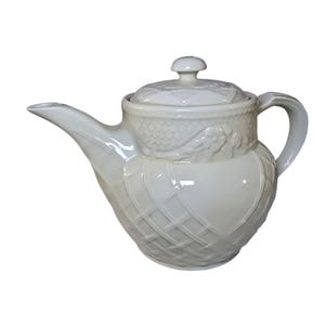 May include: A white ceramic teapot with a lid and handle. The teapot features a textured, basketweave-like pattern on the body and a decorative design around the middle. The spout is long and curved.