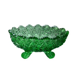 May include: A vibrant emerald green glass bowl featuring a detailed pressed starburst and diamond pattern across its exterior surface. The bowl has a scalloped rim and is supported by three small, curved feet that elevate the circular vessel.