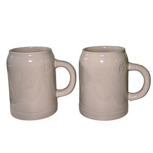 Puede incluir: Dos jarras de cerveza de cerámica beige con asas grandes. Las jarras tienen una forma cilíndrica clásica con una base ligeramente cónica. Están hechas de un material cerámico brillante y tienen un diseño simple y elegante.