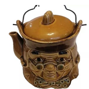 May include: A brown ceramic teapot with a lid and a handle. The teapot is shaped like a person's head with a brown hat, glasses, and a bow tie.