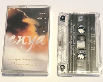 Könnte beinhalten: Ein transparentes Plastik-Kassettengehäuse mit einem silbernen Kassettenband im Inneren. Das Kassettenband trägt den Text "enya". Das Gehäuse trägt den Text "neoera performs a tribute to enya a unique interpretation of the essence of enya".