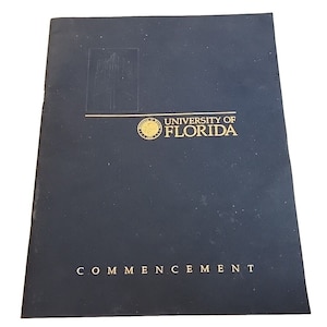 Op de afbeelding: Een donkerblauw programmaboekje voor een afstudeerceremonie van de University of Florida. De omslag toont het zegel van de universiteit en de woorden "University of Florida" en "Commencement" in gouden letters.