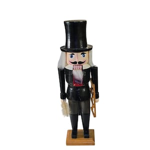 May include: A wooden nutcracker figurine in a black top hat and coat, with a white wig and mustache. The figurine holds a feather duster and a wooden ladder. The base is a light brown color.